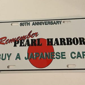May include: White license plate with black border. Text reads "50th Anniversary, Remember Pearl Harbor, Buy a Japanese Car." Features a red circle, representing the Japanese flag. Includes mounting holes.