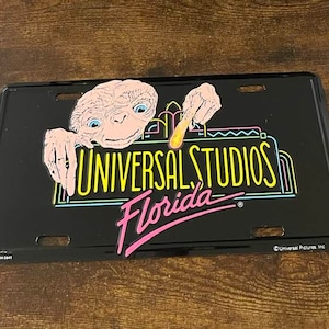 May include: A black metal license plate featuring a cartoon alien character from the movie E.T. The Extra-Terrestrial. The alien is holding a glowing stick and is peeking over a neon sign that reads "Universal Studios Florida".