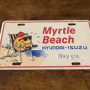 May include: White metal license plate with red and blue text that reads "Myrtle Beach Hyundai-Isuzu Hwy 501". A cartoon character wearing sunglasses and a red hat is sitting in a beach chair on a beach.