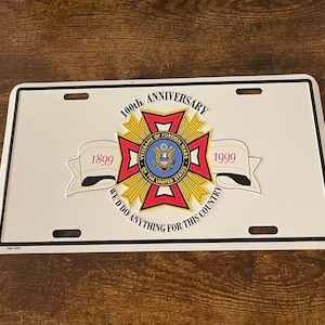 Puede incluir: Una placa de matr&iacute;cula de metal blanco con un emblema rojo, blanco y azul de los Veteranos de Guerras Extranjeras. El emblema es una cruz con una estrella en el centro. El texto "100&ordm; Aniversario" est&aacute; sobre el emblema, y los textos "1899" y "1999" est&aacute;n a ambos lados del emblema. El texto "Har&iacute;amos cualquier cosa por este pa&iacute;s" est&aacute; debajo del emblema.