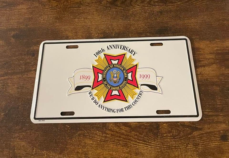 Puede incluir: Una placa de matr&iacute;cula de metal blanco con un emblema rojo, blanco y azul de los Veteranos de Guerras Extranjeras. El emblema es una cruz con una estrella en el centro. El texto "100&ordm; Aniversario" est&aacute; sobre el emblema, y los textos "1899" y "1999" est&aacute;n a ambos lados del emblema. El texto "Har&iacute;amos cualquier cosa por este pa&iacute;s" est&aacute; debajo del emblema.