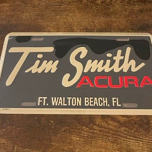 May include: A gray metal license plate with gold lettering that reads "Tim Smith Acura" and "Ft. Walton Beach, FL".