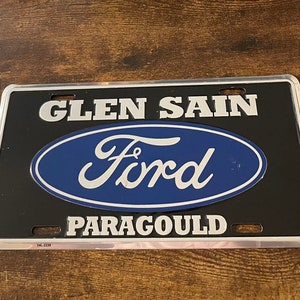 May include: A black and silver metal license plate with the text "GLEN SAIN" and "PARAGOULD" in silver lettering. A blue oval with the word "Ford" in white lettering is centered on the plate.