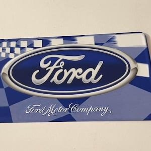 May include: A rectangular sign with the Ford logo. The logo features the word "Ford" in a silver oval against a blue background. The background has a blue and white chequered pattern. The text "Ford Motor Company" is below the logo.