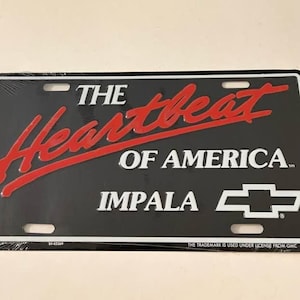 May include: A rectangular black license plate featuring the red script text The Heartbeat, white text OF AMERICA, the word IMPALA in white, and a white Chevrolet bowtie logo. The plate has four mounting holes and a white border trim.