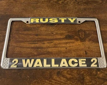 May include: A silver metal licence plate frame with black and yellow lettering. The top line reads "RUSTY" and the bottom line reads "2 WALLACE 2".