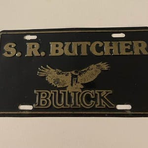 Puede incluir: Placa de matrícula negra con letras doradas y un emblema de águila dorada. El texto dice "S.R. BUTCHER" encima del águila y "BUICK" debajo. La placa tiene cuatro orificios de montaje y un borde dorado.
