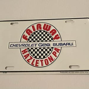 Puede incluir: Placa de matrícula blanca con borde negro. La placa presenta un círculo a cuadros rojo y negro con las palabras "FAIRWAY" y "HAZLETON, PA" en rojo. Las palabras "CHEVROLET Geo SUBARU" están impresas en negro.