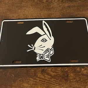 May include: Black license plate with a white rabbit head logo. The rabbit is depicted in profile, wearing a bow tie. The plate has a silver border and four mounting holes.