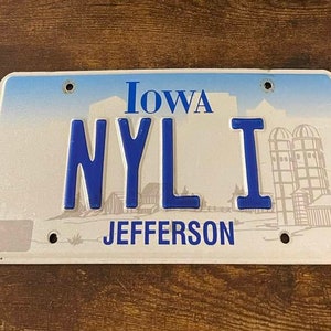 Op de afbeelding: Een wit kenteken met blauwe tekst die "Iowa NYLI JEFFERSON" luidt. De plaat heeft een blauwe lucht en een tekening van graansilo's.
