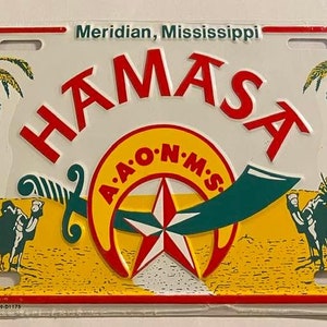 May include: A white metal license plate with the text "HAMASA" in red letters and "Meridian, Mississippi" in black letters. The plate features a yellow and green design with a star and a scimitar.