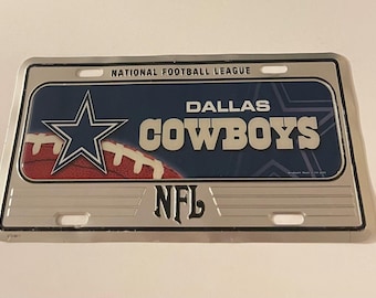 May include: A silver metal Dallas Cowboys NFL license plate with a blue and white star logo and a football. The text "National Football League" and "Dallas Cowboys" is printed on the plate.