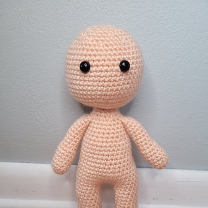 May include: A crocheted doll with a peach-coloured body and black eyes. The doll is standing on a wooden floor.