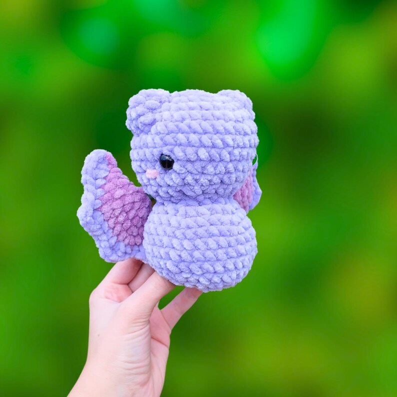 K&ouml;nnte beinhalten: Ein geh&auml;keltes lila und pinkfarbenes Fledermaus-Pl&uuml;schtier mit gro&szlig;en Augen. Das Spielzeug wird von einer Hand vor einem gr&uuml;nen Hintergrund gehalten.