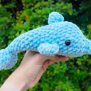 Puede incluir: Un juguete de peluche de delf&iacute;n de crochet azul claro con ojos negros. El delf&iacute;n est&aacute; en la mano de una persona.