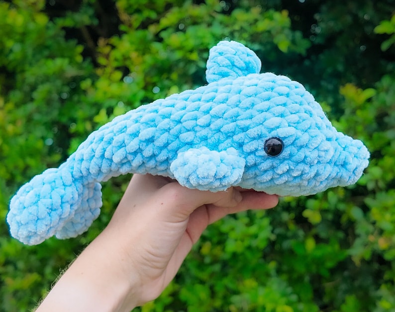 Puede incluir: Un juguete de peluche de delf&iacute;n de crochet azul claro con ojos negros. El delf&iacute;n est&aacute; en la mano de una persona.