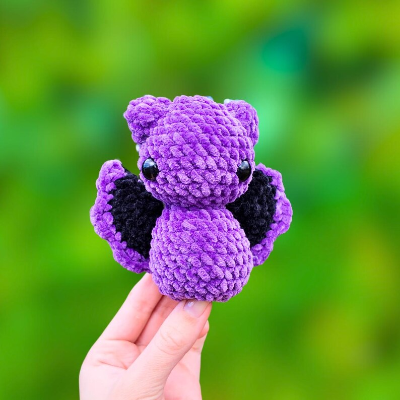 K&ouml;nnte beinhalten: Eine kleine, lilafarbene geh&auml;kelte Fledermaus mit schwarzen Fl&uuml;geln. Die Fledermaus hat gro&szlig;e schwarze Augen und eine flauschige Textur.