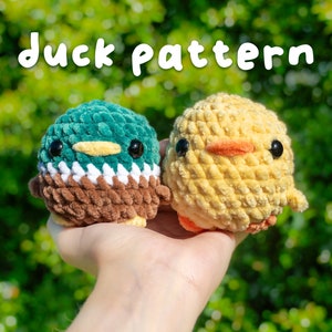 May include: Two crocheted stuffed animals, a duck and a chick, are held in a hand against a green background. The duck is green, brown, and white, and the chick is yellow. The text "duck pattern" is written in white letters above the animals.