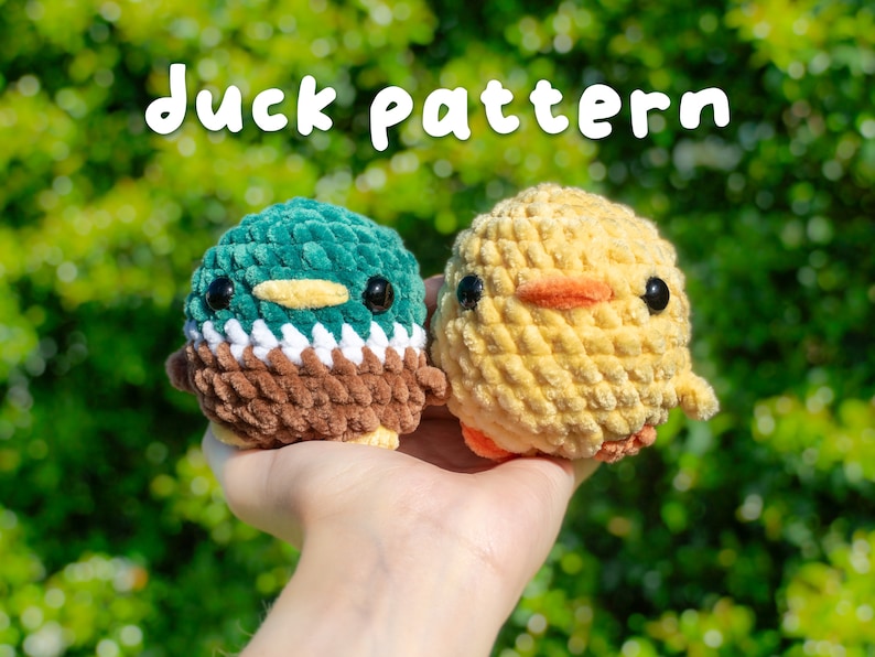 May include: Two crocheted stuffed animals, a duck and a chick, are held in a hand against a green background. The duck is green, brown, and white, and the chick is yellow. The text "duck pattern" is written in white letters above the animals.
