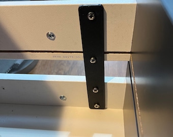 May include: Close-up of a black metal bracket attached to a white drawer with four screws. The bracket is secured to the drawer with screws. The drawer is labeled with the text "3595 22177 00.0".
