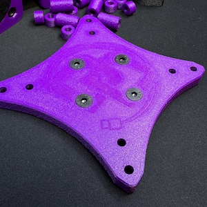 May include: A vibrant purple, four-pointed object with multiple holes and black fasteners. The object is surrounded by smaller purple cylindrical pieces and other components, all on a dark surface.