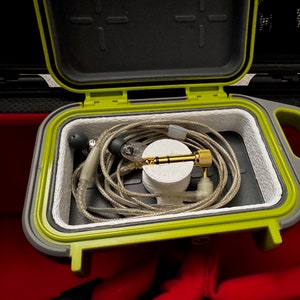 May include: A gray and green waterproof case, open to reveal in-ear monitors and a gold-colored audio jack. The case has a lime green seal and a handle. The earphones are coiled inside the case.