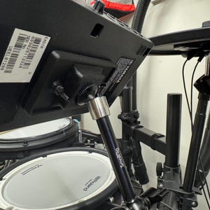 May include: A black electronic drum kit with a Yamaha module and Roland drum pads. The kit includes a SmallRig arm and a cymbal. The image shows the drum kit from a low angle, highlighting the various components and their arrangement.