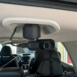 May include: A black dashcam mounted to the ceiling of a car with a white mounting plate that says "MAX 3kg/6lbs". The camera is pointed towards the back seat.