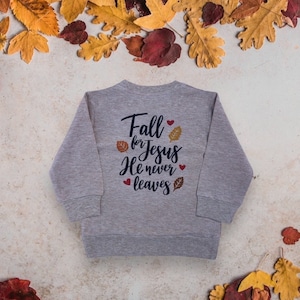 Könnte beinhalten: Grauer Pullover mit dem Text "Fall for Jesus He never leaves" in schwarzer Schrift mit kleinen roten Herzen und Blattmotiven.