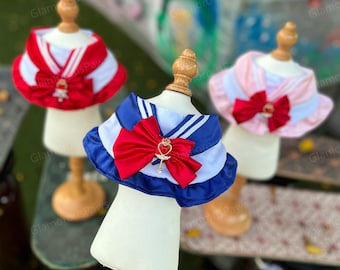 Pode incluir: Três fantasias para animais de estimação com golas estilo marinheiro em vermelho, azul e rosa. Cada fantasia tem um grande laço e enfeites decorativos. As fantasias são exibidas em pequenos manequins.