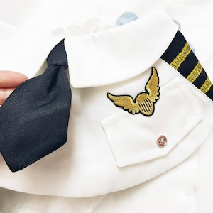 May include: White pet pilot uniform with a black tie, gold embroidered wings, and a pocket with a silver button. The collar has black and gold stripes. The text "GlamordusPaws" is visible.