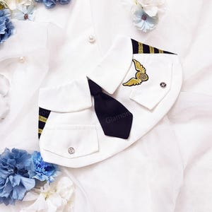May include: A white pet costume resembling a pilot's uniform, featuring a black tie, gold wing emblem, and gold stripes on the collar. The costume includes a pocket with a decorative button. The word "Glamore" is visible on the tie.