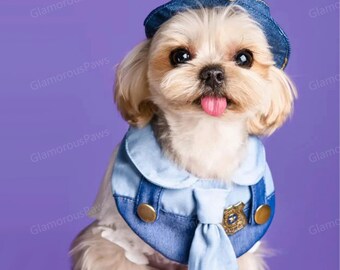 Pode incluir: Um cão pequeno e de cor clara usando um chapéu e uniforme de policial de ganga. O uniforme do cão inclui uma gola azul com botões dourados, um distintivo e uma gravata azul clara. O cão está com a língua de fora.