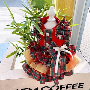 May include: A festive red and green plaid dog dress with gold accents. The dress features a ruffled skirt, a white bow, and decorative buttons. The dress is displayed on a white mannequin.