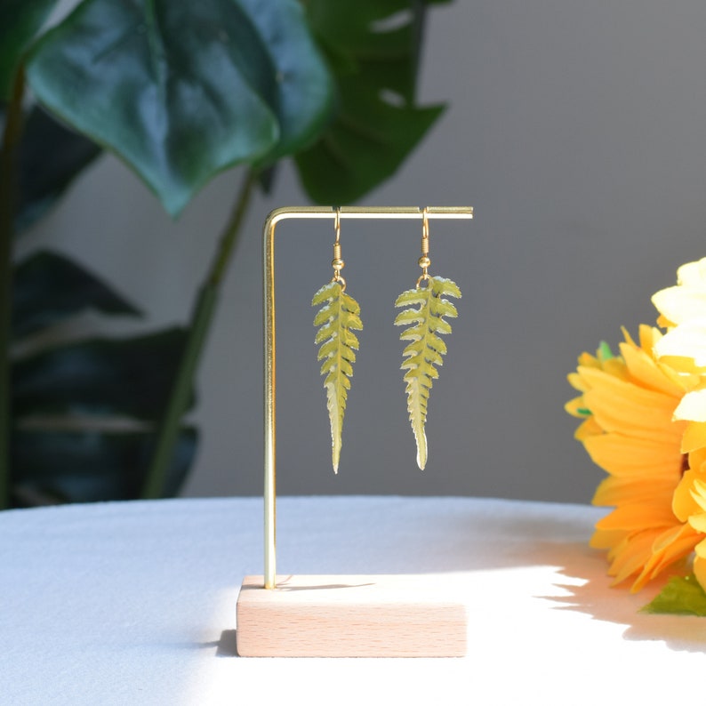 Op de afbeelding: Een paar gouden oorbellen met groene varenbladontwerpen. De oorbellen worden getoond op een houten standaard.