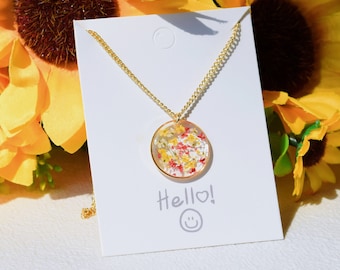 Puede incluir: Un collar dorado con un colgante circular lleno de pequeñas flores secas de colores. El colgante está enmarcado en oro y cuelga de una delicada cadena. La tarjeta detrás del collar dice "Hello!" con una carita sonriente.