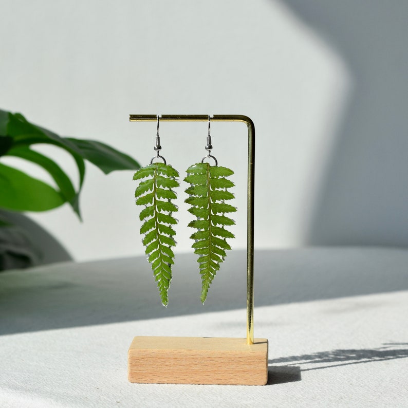Op de afbeelding: Een paar groene varenblaadjes oorbellen op een houten standaard. De oorbellen zijn gemaakt van hars en hebben een zilveren haak.