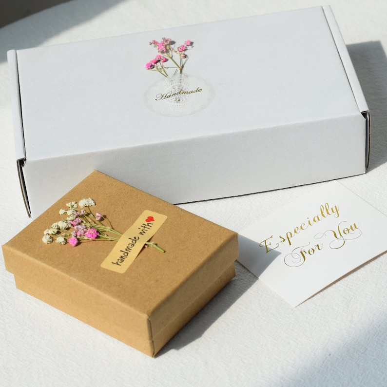 Op de afbeelding: Een klein bruin doosje met een gedroogde bloemenarrangement in wit en roze en een sticker met de tekst "handgemaakt met liefde". Het doosje staat op een wit oppervlak naast een wit doosje met een sticker met de tekst "Handgemaakt" en een witte kaart met gouden letters die "Speciaal voor jou" zegt.