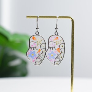 May include: A pair of silver earrings with a face outline design. The earrings are filled with pressed flowers in shades of pink, blue, orange, and white.