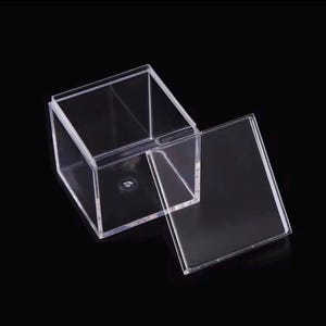 May include: A clear acrylic box with a removable lid, set against a black background. The box is cube-shaped, with the lid slightly ajar, revealing the interior. The transparent material allows a view through the sides.