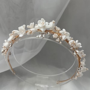 May include: A white floral headband with gold wire and pearl accents. The headband is on a clear display stand.