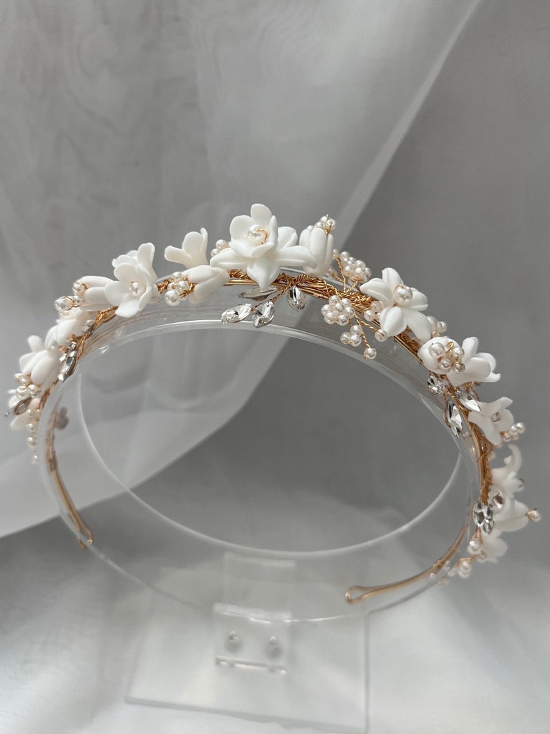 May include: A white floral headband with gold wire and pearl accents. The headband is on a clear display stand.