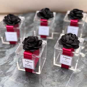 May include: Clear acrylic boxes with black fabric flowers on top. Each box is wrapped with a magenta ribbon and a white label with the names "Ashlynn & Caden" and the date "06.13.2026".