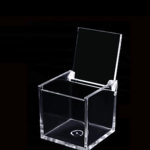 May include: A clear acrylic box with a hinged lid. The box is empty and has a square shape.