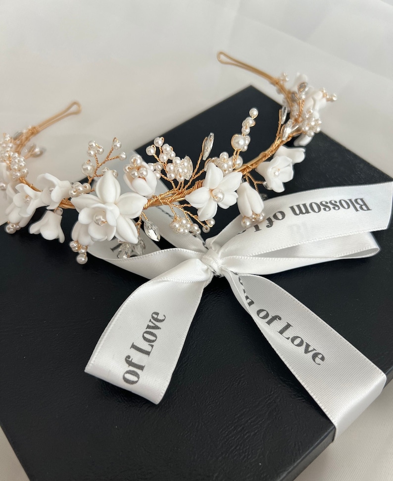 May include: A gold headband with white flowers and pearls. The headband is tied with a white ribbon that says "Blossom of Love".