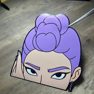 May include: A die-cut sticker featuring a stylized illustration of a woman's face. The woman has purple hair styled in a bun, with dark eyebrows and eyes. She is wearing a black earring. The sticker is on a wooden surface.