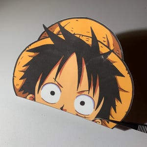 May include: A die-cut vinyl sticker featuring a cartoon character with black hair and a large yellow straw hat. The character has wide, circular eyes and a small scar under the left eye. The sticker is being held by a pair of black metal tweezers.