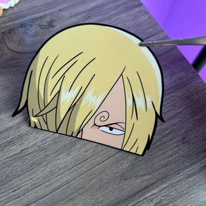 May include: A die-cut vinyl sticker featuring a stylized anime character head with blonde hair, a single visible eye with a dark eyebrow, and a distinct swirl pattern on the brow. The sticker has a black outline and a smooth, glossy finish.