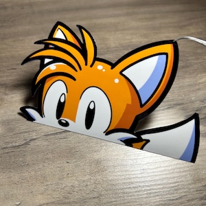 May include: A die-cut sticker featuring the character Tails from the Sonic the Hedgehog video game series. The sticker depicts Tails' orange and white head and tail, with black outlines and blue accents. The sticker is on a wooden surface.