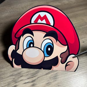May include: A red and black cartoon character with a mustache and a red hat with a white 'M' on it. The character is looking to the right and has a surprised expression.